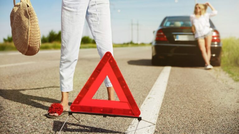 Scène de panne sur une route ensoleillée, avec un triangle de signalisation au premier plan et une femme adossée à une voiture noire en arrière-plan, semblant attendre de l’aide.