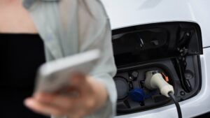 Une femme utilise son smartphone en attendant que sa voiture électrique se recharge, avec le câble branché sur la borne de recharge.