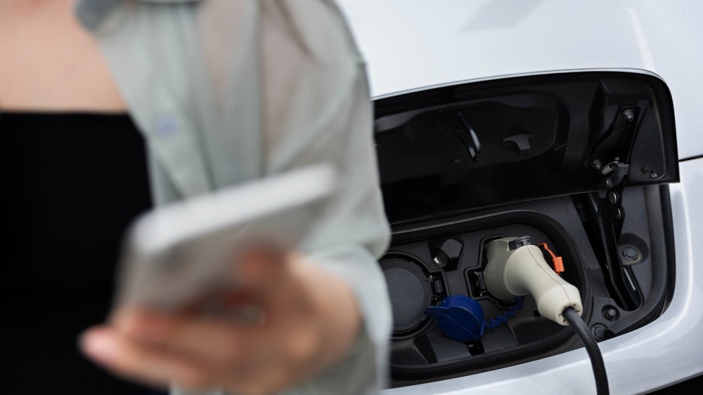 Une femme utilise son smartphone en attendant que sa voiture électrique se recharge, avec le câble branché sur la borne de recharge.